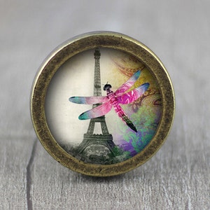 May include: A round, brass-coloured cabinet knob with a glass insert featuring a pink and purple dragonfly in front of the Eiffel Tower. The background is a faded, vintage-style design.