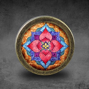 Puede incluir: Un pomo redondo de color latón con un diseño de mandala colorido. El mandala presenta una flor rosa con acentos azules y morados sobre un fondo amarillo.