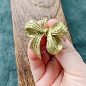 May include: A gold-colored bow brooch with detailed textures. The bow design features multiple loops and a central knot. The brooch is likely made of metal and is a decorative accessory.