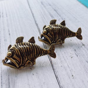 Könnte beinhalten: Zwei goldfarbene Fischfiguren aus Metall mit detaillierten Schuppen und offenem Maul. Die Fische sind in die gleiche Richtung ausgerichtet und befinden sich auf einer weißen Holzoberfläche.