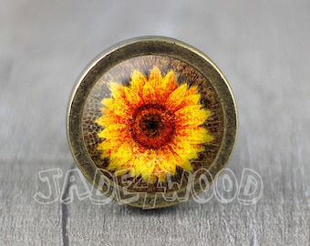 Può includere: Pomello per armadio rotondo color bronzo con un motivo a girasole. Il girasole ha petali gialli con accenti rossi e un centro marrone scuro. Il testo "JADE4WOOD" è visibile sulla superficie.