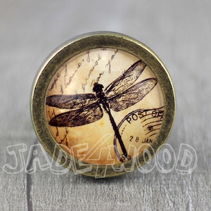 May include: A round, brass-coloured cabinet knob with a glass insert featuring a vintage-style dragonfly illustration on a cream-coloured background with handwritten text and a "POST OFFICE" stamp. The stamp reads "28 JAN".