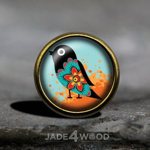 Puede incluir: Un botón redondo de metal dorado con un diseño de pájaro colorido. El pájaro es negro con una flor azul, turquesa y roja en el pecho. El fondo es azul claro con salpicaduras naranjas.