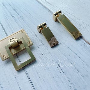 May include: A set of gold-coloured cabinet handles. The set includes a rectangular pull with a square handle and two rectangular pulls with wave patterns. The hardware is displayed on a light-coloured wooden surface.