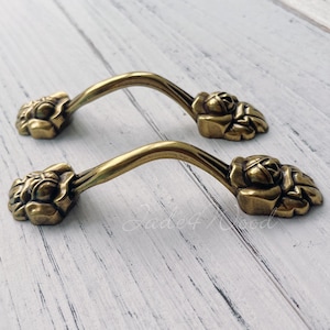 May include: Two ornate brass drawer pulls with a floral design. The pulls are curved and have a decorative, raised floral pattern.