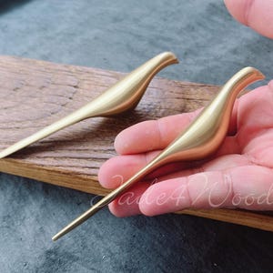 May include: Two gold metal bird-shaped hooks. The hooks are on a wooden surface and a hand is holding one of the hooks.