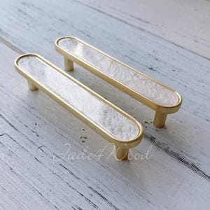 Puede incluir: Dos tiradores de armario con acabado dorado e incrustaciones de mármol blanco. Los tiradores tienen forma rectangular con extremos redondeados y un pequeño tirador cilíndrico. Herrajes para muebles y decoración del hogar.