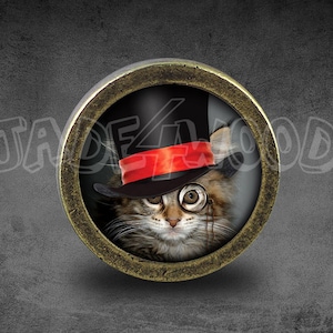 Puede incluir: Un marco redondo de color latón con un inserto de vidrio. Dentro del marco hay una imagen de un gato que lleva un sombrero de copa negro con una banda roja y un monóculo.