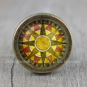 May include: A round, brass-colored cabinet knob with a colorful geometric design. The design features a starburst pattern with red, yellow, and brown colors.