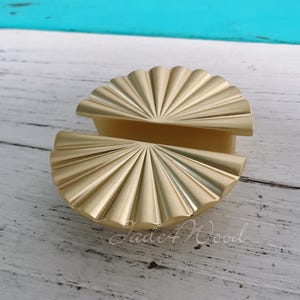 Pode incluir: Uma maçaneta de armário de metal dourado com um design em forma de leque. A maçaneta é meio círculo e tem uma superfície texturizada em relevo.