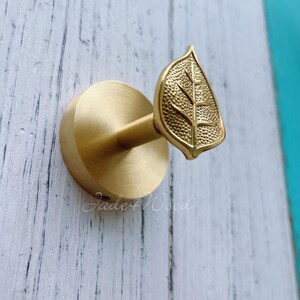 May include: A brass leaf-shaped hook with a round base, mounted on a white wooden surface. The hook features a detailed leaf design and the text "Jade4Wood". Ideal for adding a touch of nature-inspired decor.