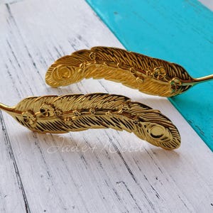 Könnte beinhalten: Zwei goldfarbene Haarspangen in Federform. Die Spangen weisen aufwendige Details auf, darunter eine zentrale Mittelrippe und zarte Federborsten. Der Hintergrund ist eine Mischung aus weiß und türkisfarben lackiertem Holz.