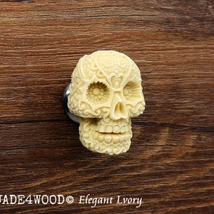 May include: Ivory-colored skull-shaped cabinet knob with intricate floral designs. The skull has a detailed face with eye sockets, a nose, and a mouth. The knob is made of a smooth, polished material.