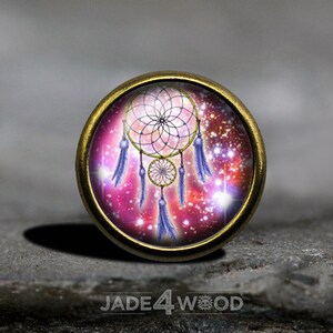 Puede incluir: Un charm redondo dorado con un fondo de galaxia rosa y morado y un atrapasueños dorado con plumas moradas.