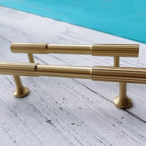 Puede incluir: Dos tiradores de armario dorados con un diseño acanalado. Los tiradores son de metal y tienen un estilo moderno.