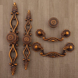 May include: An assortment of antique bronze cabinet hardware, including two long pulls, two smaller pulls, and a round knob. The hardware features floral designs and ornate detailing, perfect for adding a vintage touch to furniture.