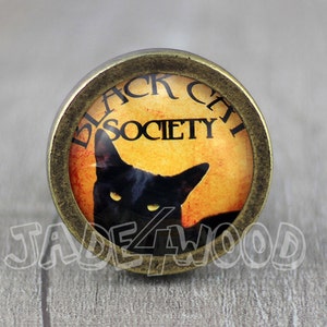 Könnte beinhalten: Ein runder, goldfarbener Knopf mit einer schwarzen Katze und den Worten "Black Cat Society" auf gelbem Hintergrund.