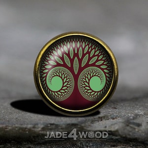 Könnte beinhalten: Ein runder, goldfarbener Metallknopf mit einem grünen und burgunderfarbenen Design. Das Design zeigt einen stilisierten Baum mit einem sich wiederholenden Muster aus Blättern.