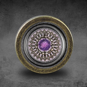 May include: A round, ornate, silver and gold ring with a purple gemstone surrounded by smaller diamonds. The ring is set in a black background.