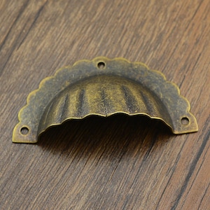 Könnte beinhalten: Ein vintage-artiger, messingfarbener Schrankgriff mit einem gewellten Rand und einem geriffelten Design. Der Griff ist für die Montage an einer Schranktür konzipiert.