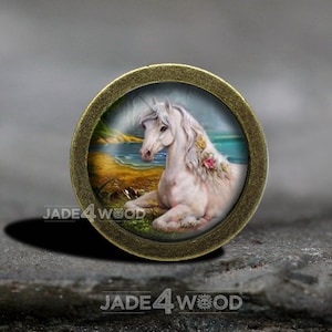 Może przedstawiać: Biały jednorożec ze złotym rogiem i kwiatami w grzywie leży na plaży z niebieskim oceanem w tle. Obraz jest osadzony w złotym metalowym kole.