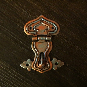 Puede incluir: Un pestillo de metal decorativo con un acabado en cobre y plata. El pestillo tiene una parte superior en forma de corazón con detalles intrincados y una cerradura en el centro.