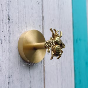 Puede incluir: Un pomo de armario con forma de abeja de color latón. El pomo presenta un diseño detallado de abeja con alas y segmentos corporales. La base redonda está unida a un vástago cilíndrico. El fondo es una superficie de madera pintada de blanco y turquesa.