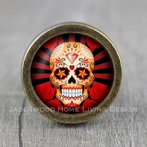 May include: A decorative cabinet knob with a Day of the Dead sugar skull design. The skull is orange and yellow with a diamond and star accents, set against a red and black striped background. Red roses surround the skull, and the knob has a bronze finish.