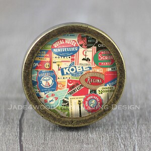 Puede incluir: Un pomo redondo de color latón con un tema de viaje vintage. El pomo está cubierto de pegatinas coloridas con texto como "Hotel Kobe", "Orient Express" y "Hotel Regina Paris".