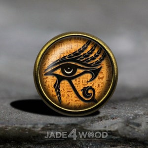 May include: A gold-toned round button featuring a black and white image of the Eye of Horus against a background of orange hieroglyphics.