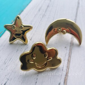 May include: Three gold-colored cabinet knobs in the shape of a smiling star, cloud, and crescent moon. The knobs have a shiny finish and are set against a white and blue wooden surface. The knobs are likely for a child's room or nursery.