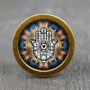 Peut inclure: Un bouton rond doré avec un motif de mandala coloré représentant une main de hamsa blanche avec un cœur au centre. La main de hamsa est un symbole de protection et de bonne chance.