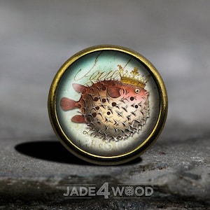 May include: A round, gold-toned metal button with a glass top featuring a whimsical illustration of a crowned pufferfish with a pink and brown body and white spines. The background is a light blue watercolour wash with gold script.