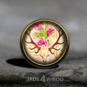 Puede incluir: Un colgante redondo dorado con una imagen de estilo vintage de un cráneo de ciervo con cuernos y rosas rosas.