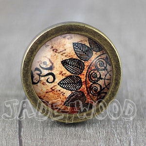 May include: A round, brass-colored cabinet knob with a glass insert. The glass insert features a vintage-inspired design with brown and black swirls, leaves, and a symbol that looks like an "Om" symbol.