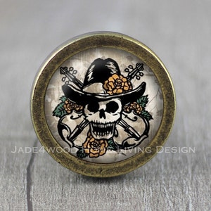 以下が含まれることがあります： カウボーイハットをかぶり、バイオリンを持った骸骨が描かれた、真鍮色の丸いノブ。骸骨は歯を見せて笑っており、花に囲まれています。ノブは家具の装飾的なアクセントです。