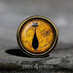 Könnte beinhalten: Ein runder, goldfarbener Metallknopf mit einer schwarzen Katzensilhouette auf gelbem Hintergrund mit einem floralen Muster. Die Katze hat weiße Augen und einen langen Schwanz.