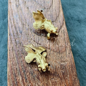 May include: Two gold-colored metal goldfish figurines on a wooden surface. The goldfish are detailed with fins, scales, and eyes.