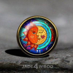 May include: A round, brass-colored pendant with a colorful design of a sun and moon with closed eyes and hearts. The sun is orange and red, the moon is yellow, and the background is blue, green, and white.
