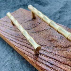 Puede incluir: Dos tiradores de cajón de estilo bambú de color dorado sobre una superficie de madera. Los tiradores tienen un acabado liso y pulido.