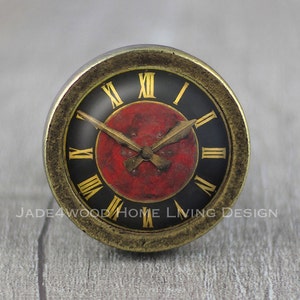 Puede incluir: Una esfera de reloj redonda de estilo antiguo con fondo negro y números romanos dorados. El centro es rojo con manecillas doradas. El reloj tiene un borde dorado. La esfera del reloj es un elemento decorativo.