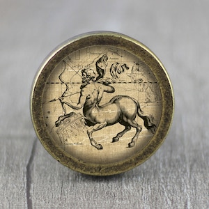 Può includere: Un pomello rotondo color ottone con un'immagine in stile vintage della costellazione del Sagittario. L'immagine è in bianco e nero e presenta un centauro che tiene un arco e una freccia.