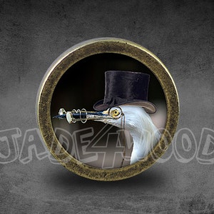Può includere: Un bottone rotondo color bronzo con un uccello bianco che indossa un cappello a cilindro nero e occhiali in stile steampunk. L'uccello è posto su uno sfondo scuro. Il testo "JADE4WOOD" è visibile.