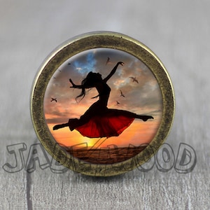 May include: A round, antique gold-toned cabinet knob with a glass insert featuring a silhouette of a woman in a red dress jumping against a sunset background with birds flying overhead.