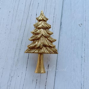 Può includere: Un ornamento dorato per l'albero di Natale. L'albero ha un design dettagliato e stratificato e un tronco sottile. L'ornamento è posto su uno sfondo chiaro con venature del legno. Il design generale è festoso e decorativo.