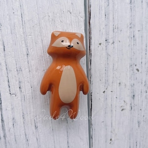 May include: A ceramic fox figurine in shades of orange and cream. The fox has a rounded body, small arms, and a friendly expression. The figurine is standing upright and has a glossy finish. The background is a white wooden surface.