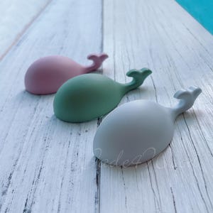 May include: Three whale-shaped ceramic hooks in pink, green, and grey. The hooks are on a white wooden surface.