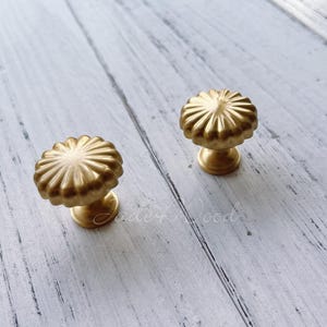 Puede incluir: Dos pomos de armario dorados con un diseño acanalado en forma de sol. Los pomos tienen una base redonda y son de metal.