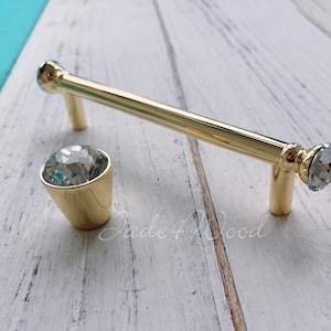 May include: A gold-coloured cabinet pull and matching knob with crystal-like accents. The pull is a bar shape, and the knob is cone-shaped. Both have a shiny finish and are set against a light wood background.