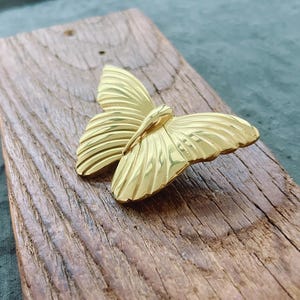 May include: A gold butterfly brooch with detailed wing patterns. The brooch is on a weathered, rectangular wooden surface. The butterfly's wings are spread, showing the intricate design. The wood has a natural grain.